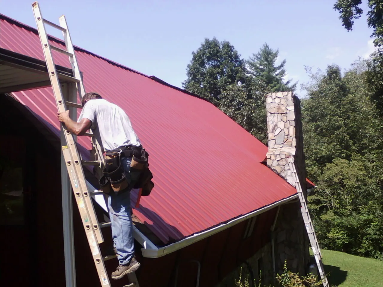 Roofer on a ladder beside a red metal roof and stone chimney — Soffit & Fascia Repair by Roofing Brothers in Rutherford